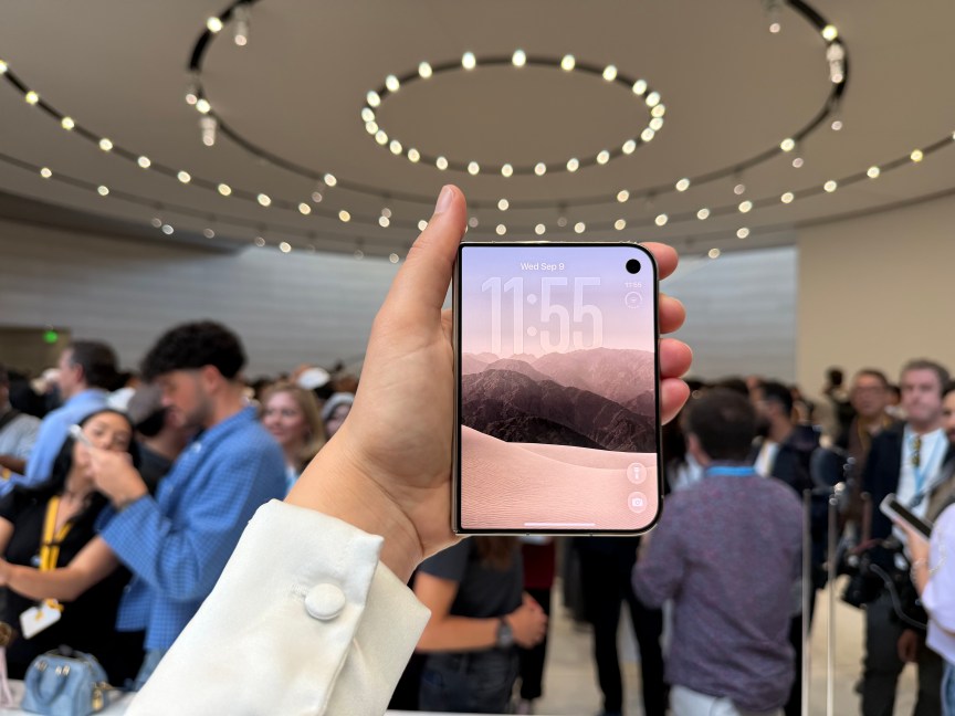 hand holding the iPhone Duo in a busy room, with the front screen showing mountains and the time 11:55